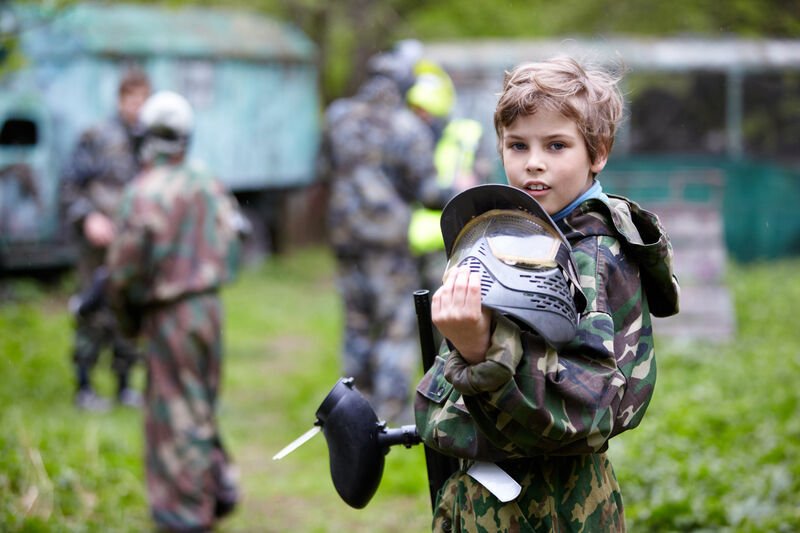 A young player holding a paintball mask and marker at a field